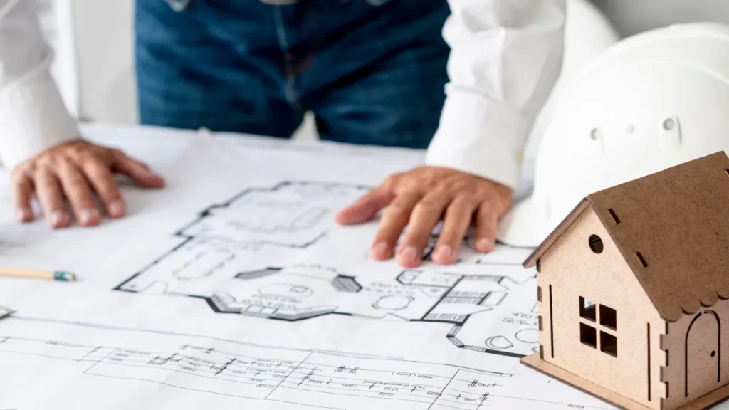 Pessoa analisando plantas arquitetônicas sobre uma mesa, com um capacete de segurança e uma maquete de casa ao lado.
