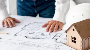 Pessoa analisando plantas arquitetônicas sobre uma mesa, com um capacete de segurança e uma maquete de casa ao lado.